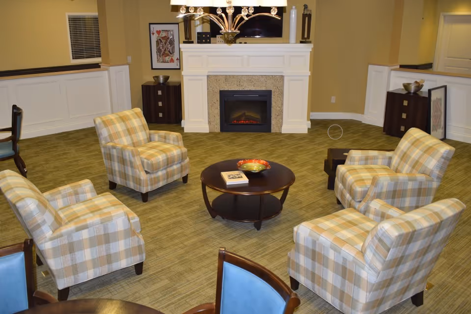 Cozy common room with four plaid armchairs arranged around a round coffee table facing a fireplace and TV.