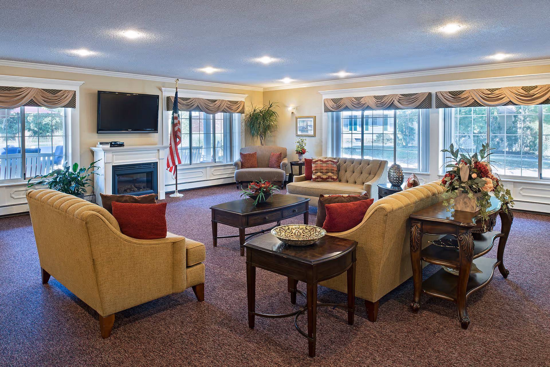 A spacious and well-lit living room area with beige sofas and armchairs arranged around a dark wooden coffee table. The room features large windows with valance curtains, a flat-screen TV mounted above a white fireplace, an American flag, and decorative plants and flowers on side tables. The carpet is a muted purple color, and the walls are painted a soft yellow.