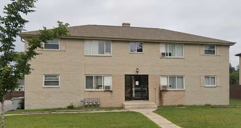 Exterior view of a two-story beige brick building with multiple windows and a central entrance door. There is a small concrete pathway leading to the entrance, and a grassy lawn in front of the building. A tree is visible on the left side of the image.