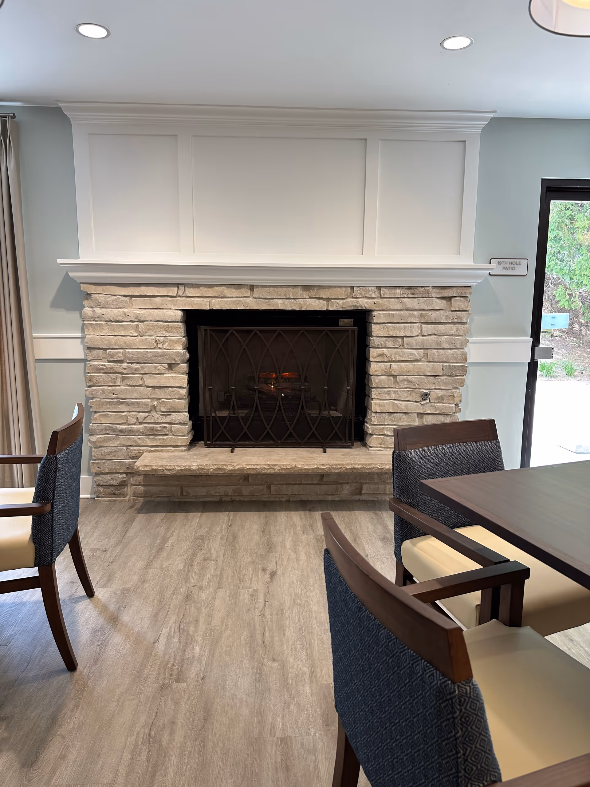 A cozy indoor space featuring a stone fireplace with a black metal screen. Surrounding the fireplace are wooden chairs with cushioned seats and backs, and a wooden table is partially visible. There is a glass door on the right side leading to an outdoor area with greenery.