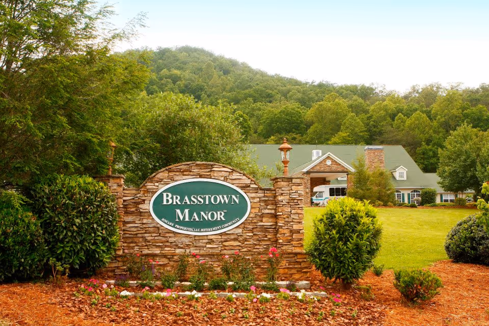 Stone sign for Brasstown Manor Senior Living in front of a landscaped lawn with bushes and flowers, with a large building and green forested hills in the background.