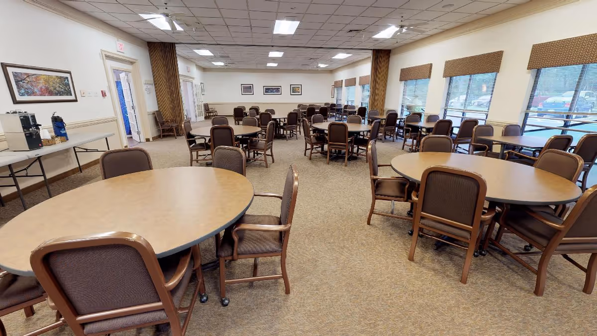 A spacious dining room in Cedar Lane Senior Living Community with multiple round tables and brown cushioned chairs arranged neatly. Large windows on the right side allow natural light to fill the room. There are framed pictures on the walls and a coffee station on a table to the left.