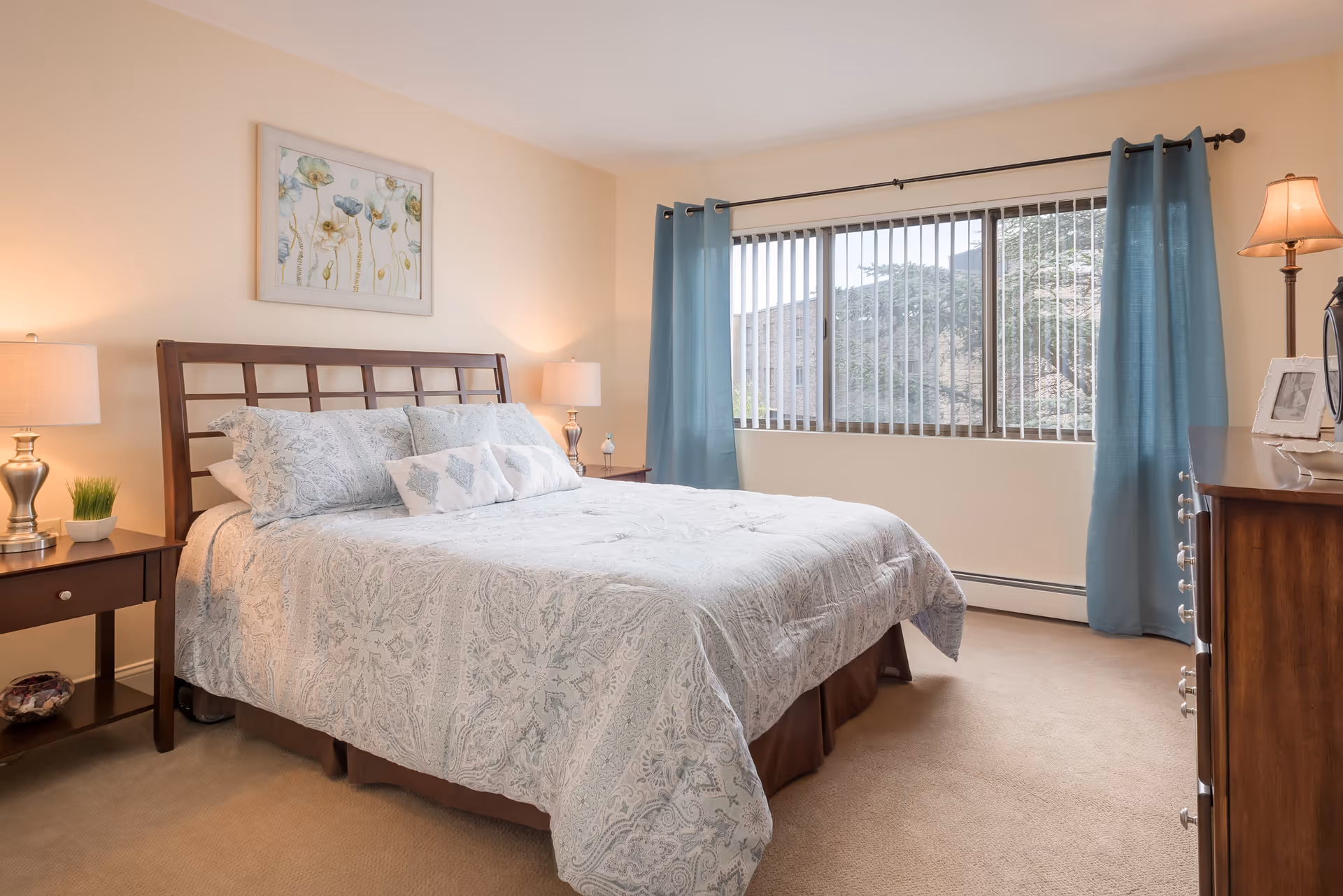 A neatly made bed with a wooden headboard in a bedroom. The bed has a light-colored patterned comforter and several pillows. On either side of the bed are wooden nightstands with lamps and small decorative items. A large window with vertical blinds and blue curtains lets in natural light. A framed floral artwork hangs on the wall above the bed, and a wooden dresser with a lamp and picture frames is visible on the right side of the room.