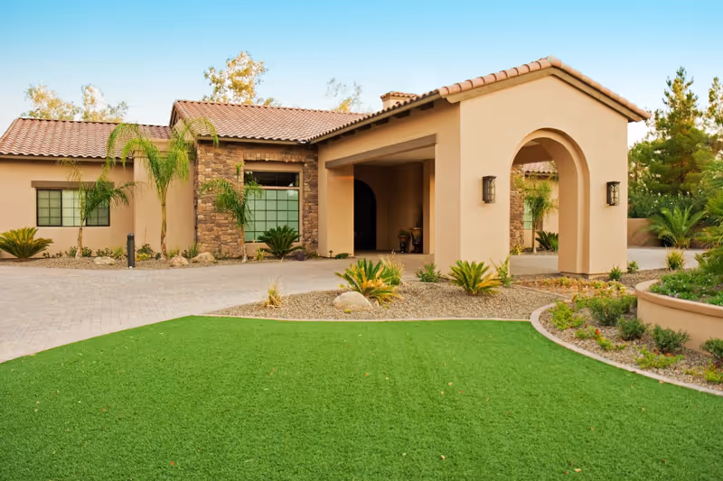 Exterior view of a single-story building with a beige stucco finish and a tiled roof, featuring an arched entrance. The surrounding landscape includes green grass, small palm trees, shrubs, and a paved driveway.