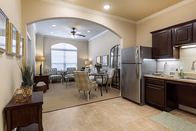 View of a senior living facility interior showing a kitchen area with dark wood cabinets, a stainless steel refrigerator, and a sink. Adjacent to the kitchen is a living room with patterned armchairs, a glass-top dining table, a ceiling fan, and a large window with blinds letting in natural light. The walls are painted beige and the floor transitions from tile in the kitchen to carpet in the living room.