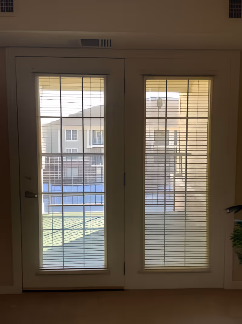 A set of white double doors with glass panels covered by horizontal blinds, leading to an outdoor area with buildings visible through the glass.