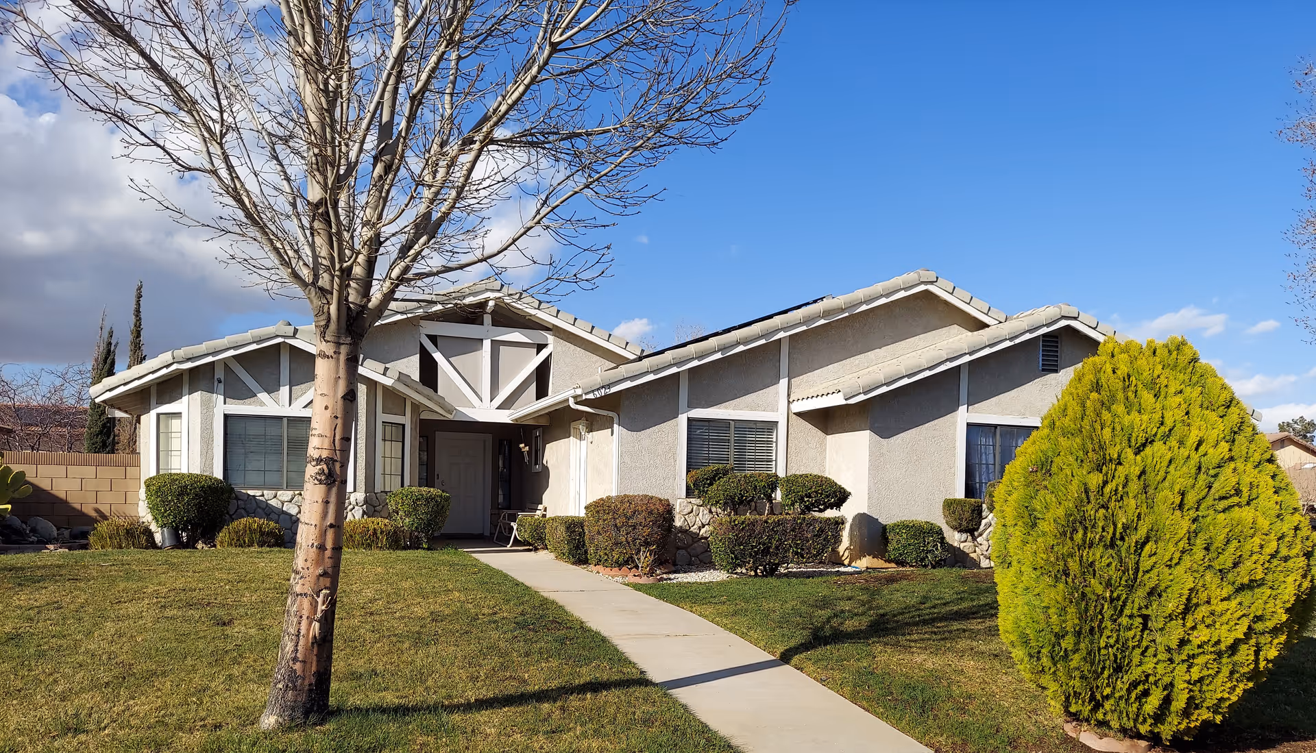 Single-story suburban house front with a walkway, lawn, trimmed shrubs and trees under a blue sky.