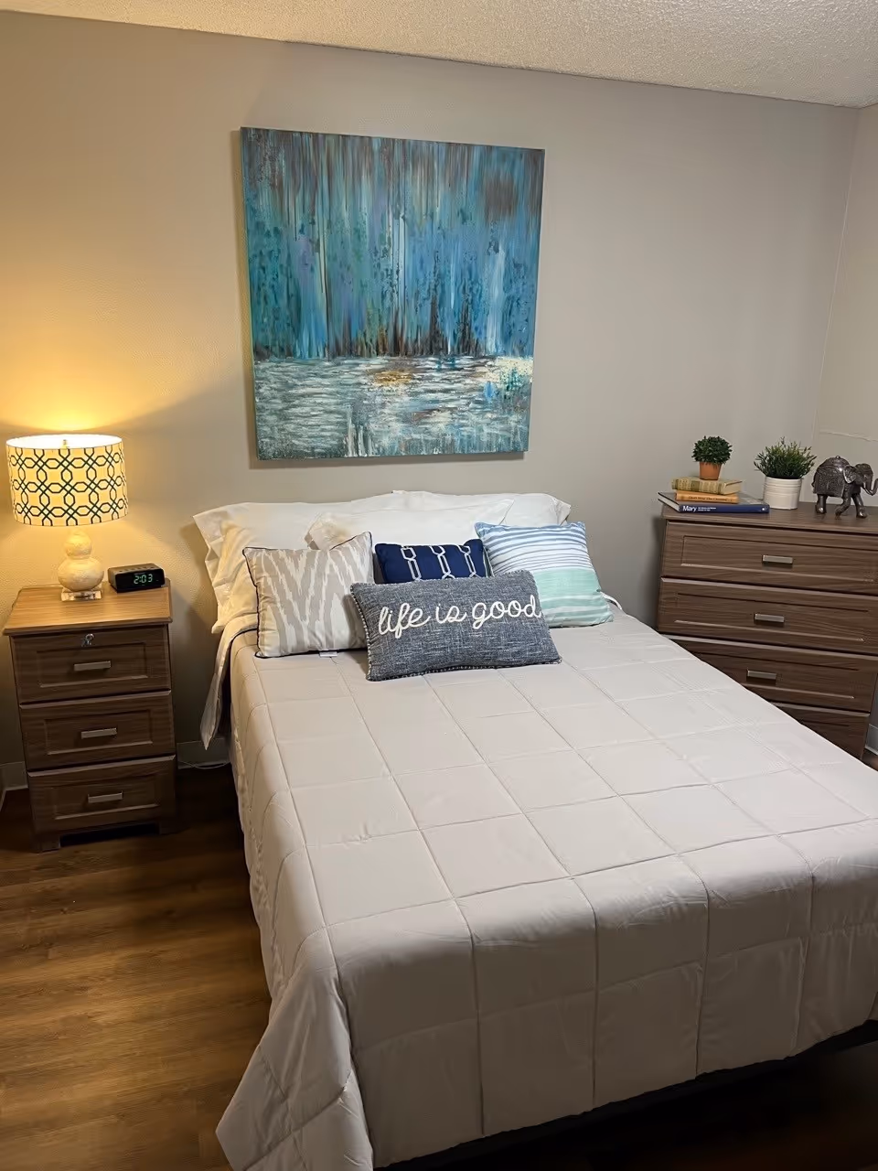 A neatly made bed with a beige quilt and multiple decorative pillows, including one with the phrase 'life is good'. On the left side of the bed is a wooden nightstand with a patterned lamp and a digital clock showing 2:03. On the right side is a wooden dresser with small potted plants, books, and an elephant figurine. Above the bed hangs a large abstract painting with blue and green tones. The room has wooden flooring and beige walls.