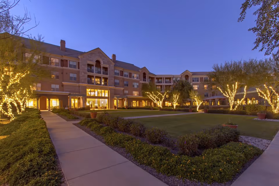 Evening view of a senior living facility named Vi at Silverstone, showing a large, multi-story building with warm interior lights visible through the windows. The landscaped courtyard features a well-maintained lawn, shrubs, and trees wrapped in decorative string lights, with paved walkways leading through the garden area.