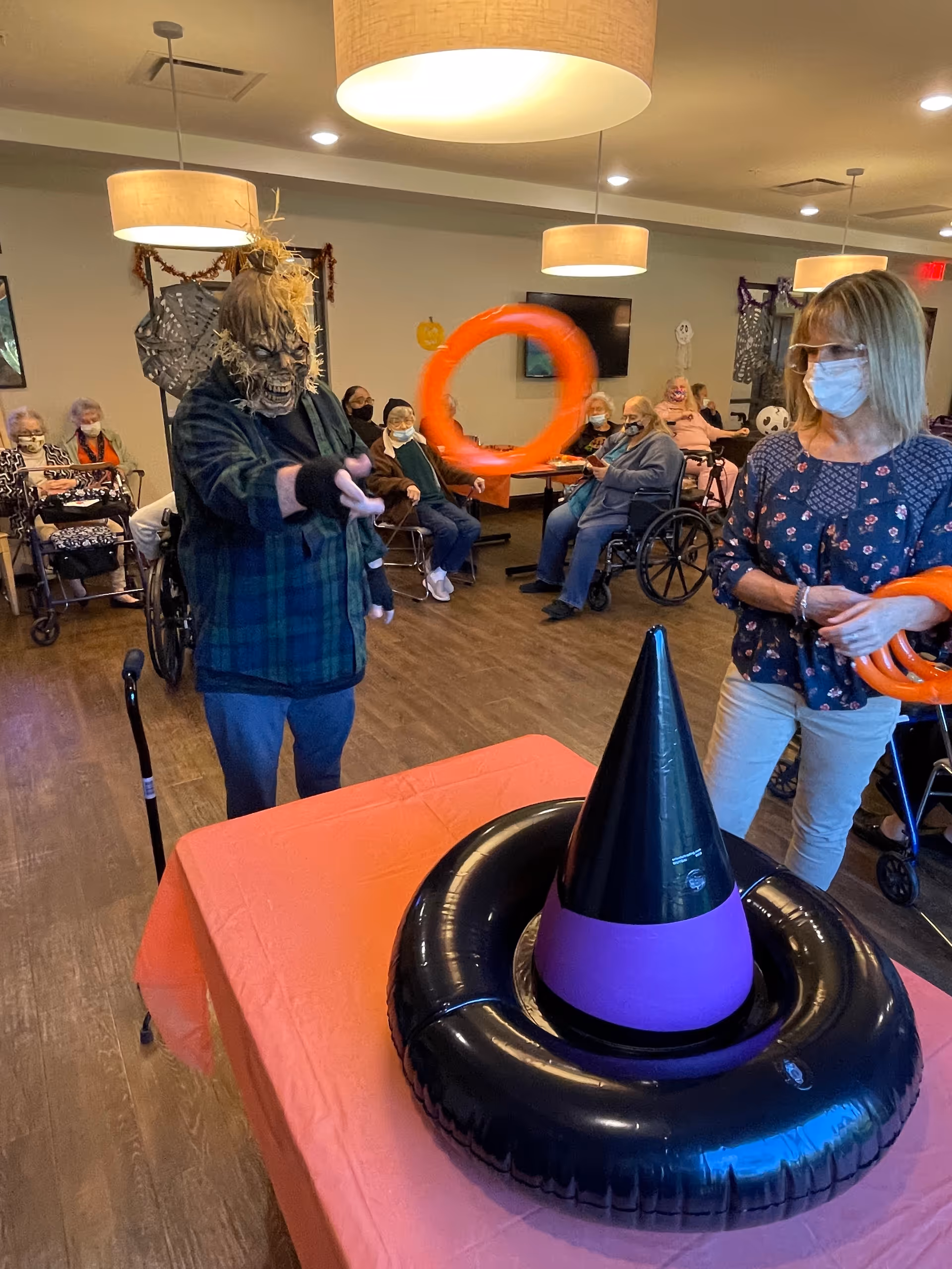 A group of elderly people, some in wheelchairs, are gathered in a decorated room. One person wearing a scary mask and plaid shirt is tossing an orange ring towards a large inflatable witch hat on a table covered with an orange tablecloth. Another person wearing a mask and glasses is holding more orange rings. The room has hanging lights and Halloween decorations on the walls.