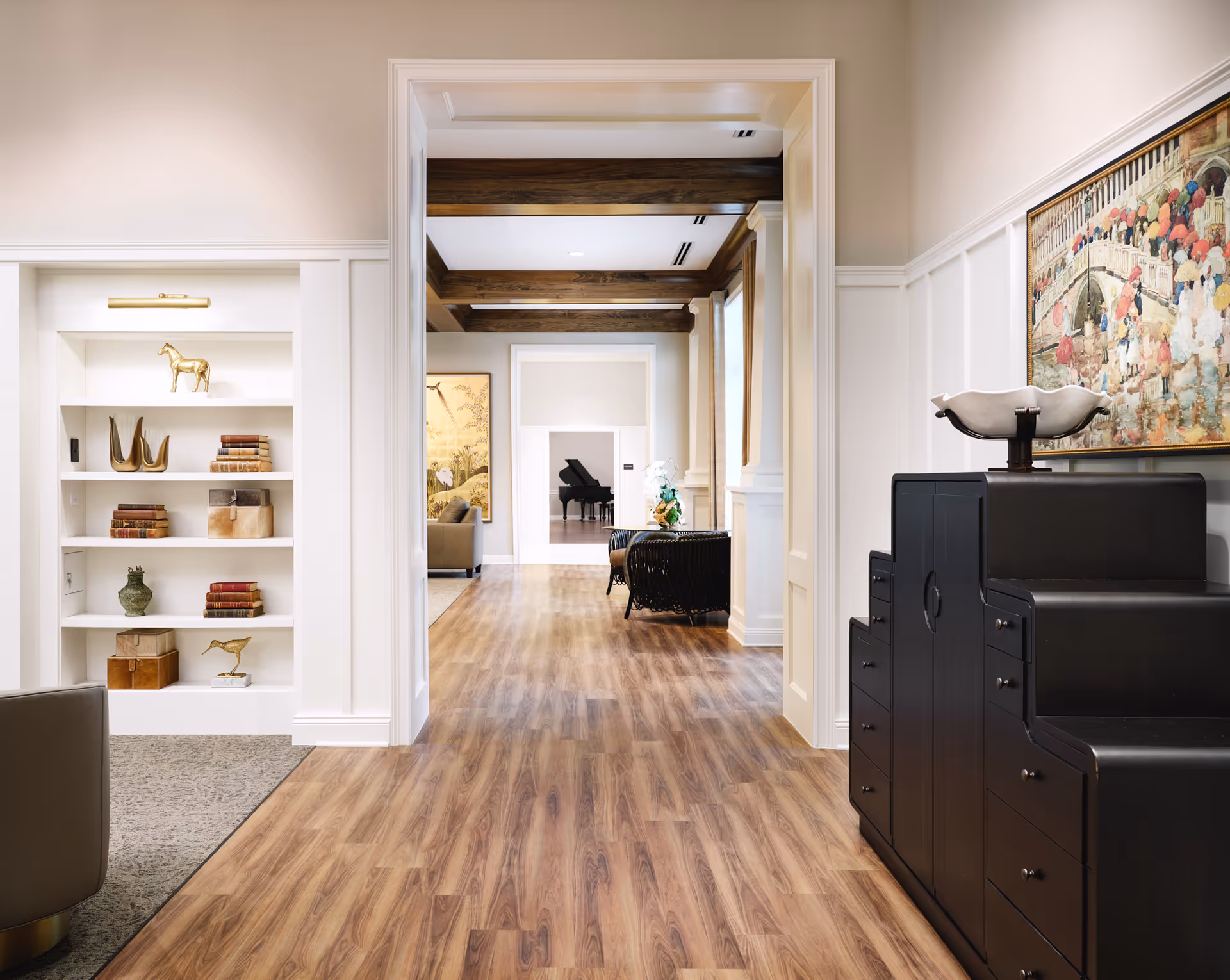 A bright hallway in a senior living facility with wood flooring and white walls. On the left, there is a built-in white bookshelf with decorative items and books. On the right, there is a black cabinet with a decorative bowl on top and a colorful painting above it. The hallway leads to a room with a grand piano and seating areas with wooden ceiling beams visible.
