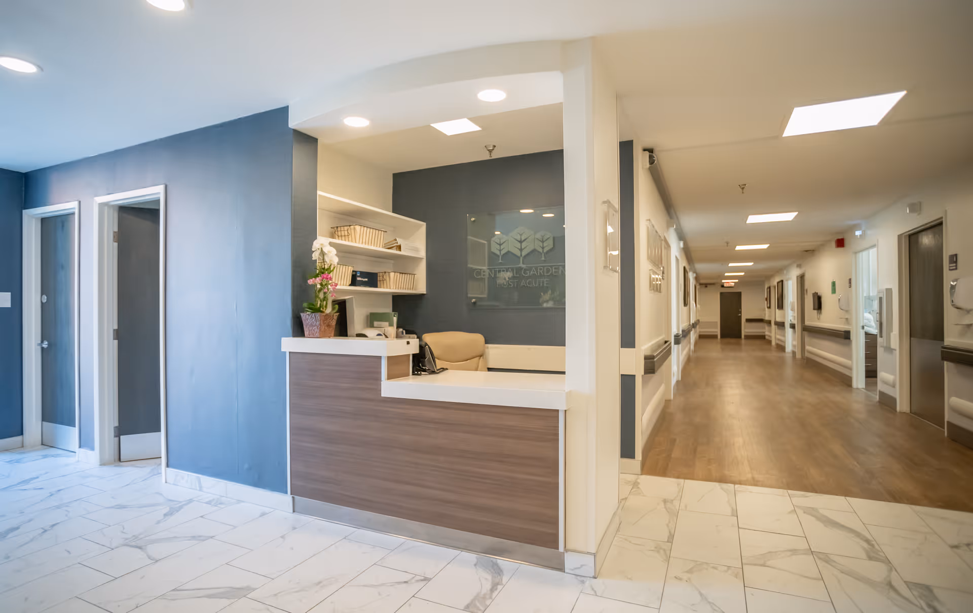 Reception desk area with a wooden counter and a beige chair behind it, located in a hallway with blue and white walls and marble flooring. The hallway extends to the right with wooden flooring and several doors along the corridor. A sign behind the reception desk reads 'Central Gardens Post Acute'.