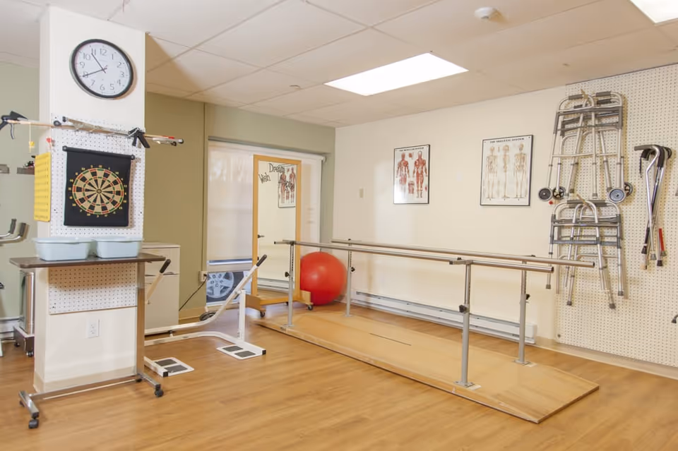 Physical therapy/exercise room with parallel bars, walkers, an exercise ball, a dartboard, and anatomical charts on the walls.