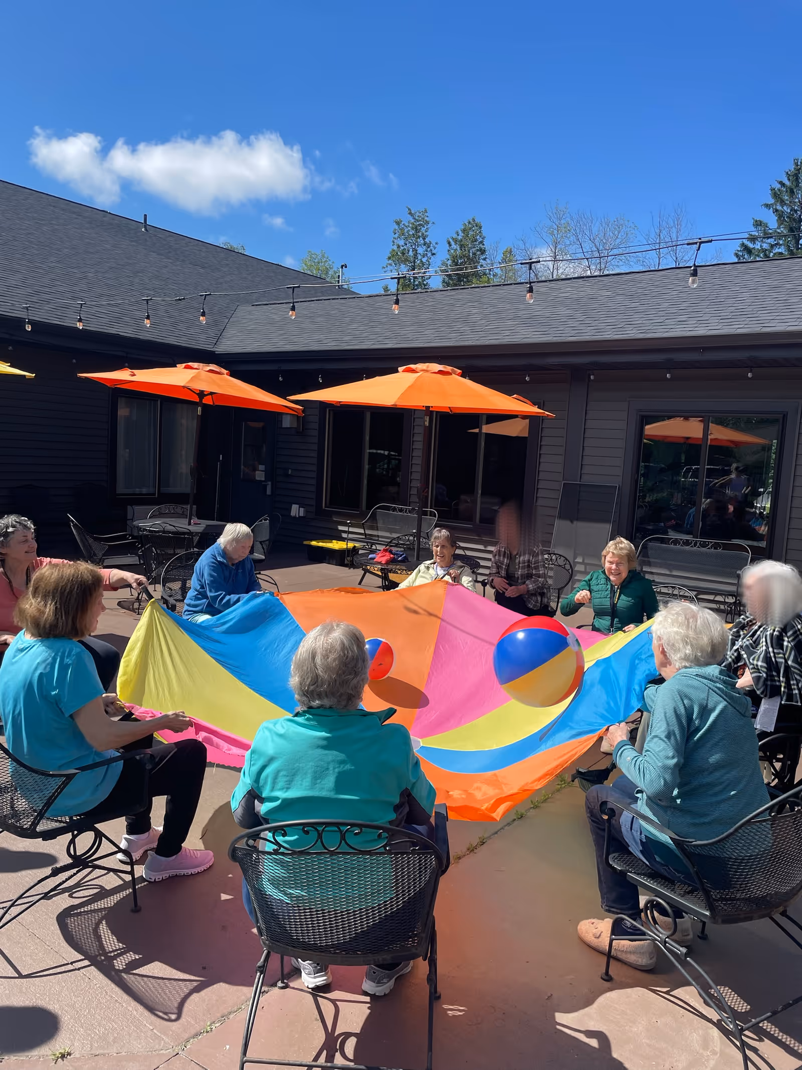 A group of elderly people sitting in a circle outdoors on a patio, holding the edges of a colorful parachute with two beach balls on top. The patio has orange umbrellas and black metal chairs, with a building in the background under a clear blue sky.