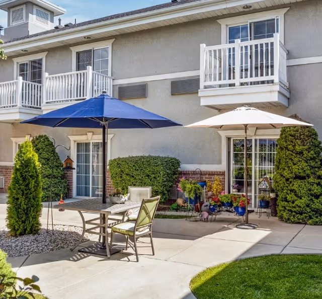 Outdoor patio area of a senior living facility with two umbrellas, one blue and one white, shading a table and chairs. The building has balconies and sliding glass doors with greenery and potted plants around the patio.