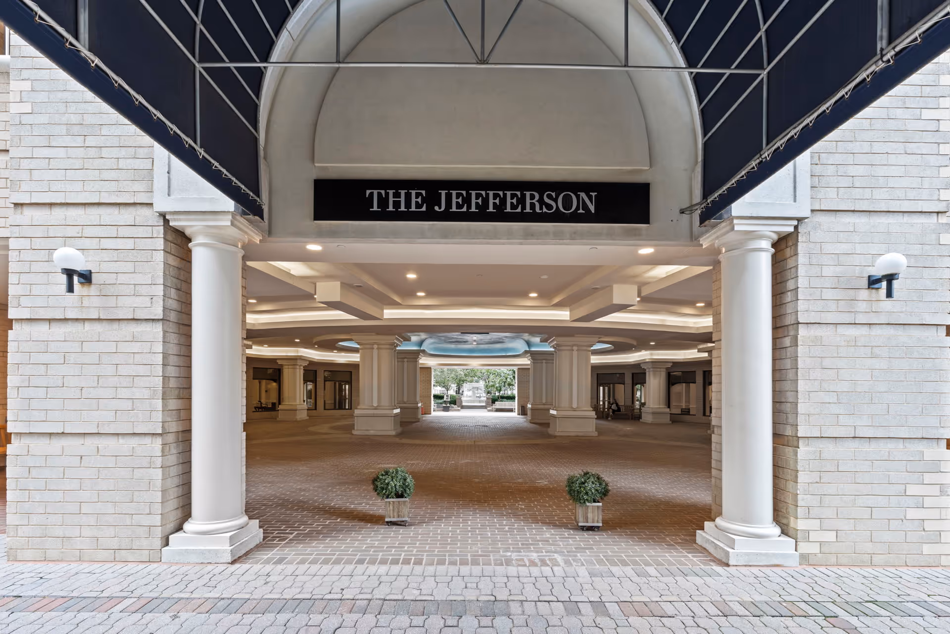 Entrance area of The Jefferson building featuring a covered driveway with white columns, brick flooring, and a black sign with white text reading 'THE JEFFERSON'. Two small potted plants are placed on the ground near the entrance.