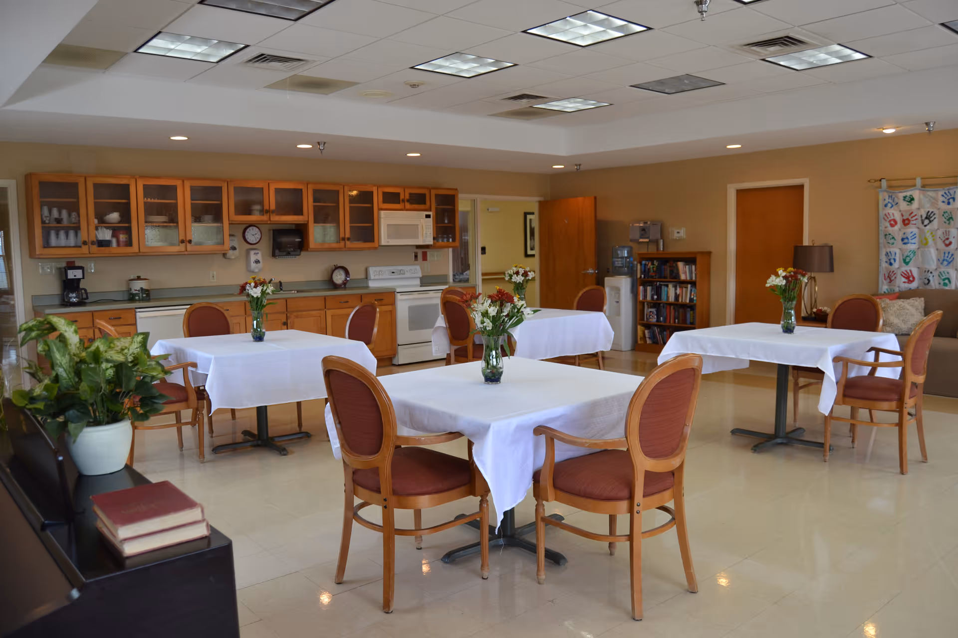 A bright and clean dining area in a senior living facility with four tables covered in white tablecloths, each adorned with a vase of flowers. The room features wooden chairs with red cushions, a kitchenette with wooden cabinets, a stove, microwave, and dishwasher in the background. There are plants, bookshelves, and a couch with a colorful handprint wall hanging on the right side.