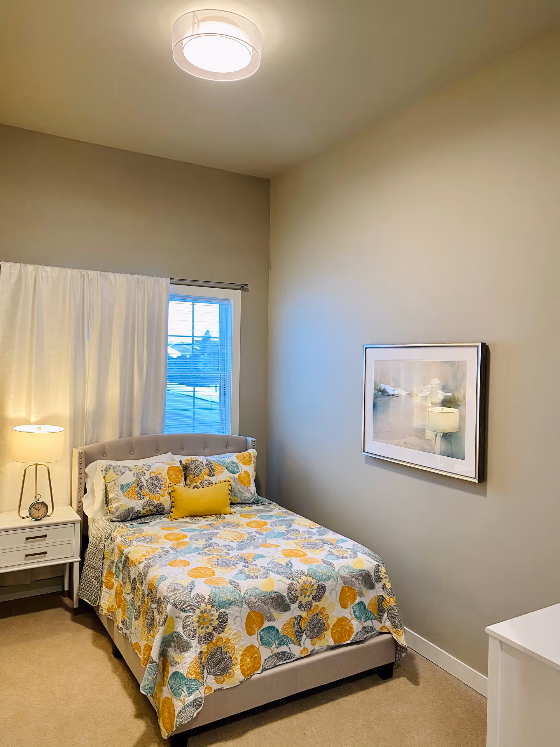 A neatly made single bed with patterned bedding, a nightstand and lamp beside a window in a softly lit bedroom with a framed picture on the wall.
