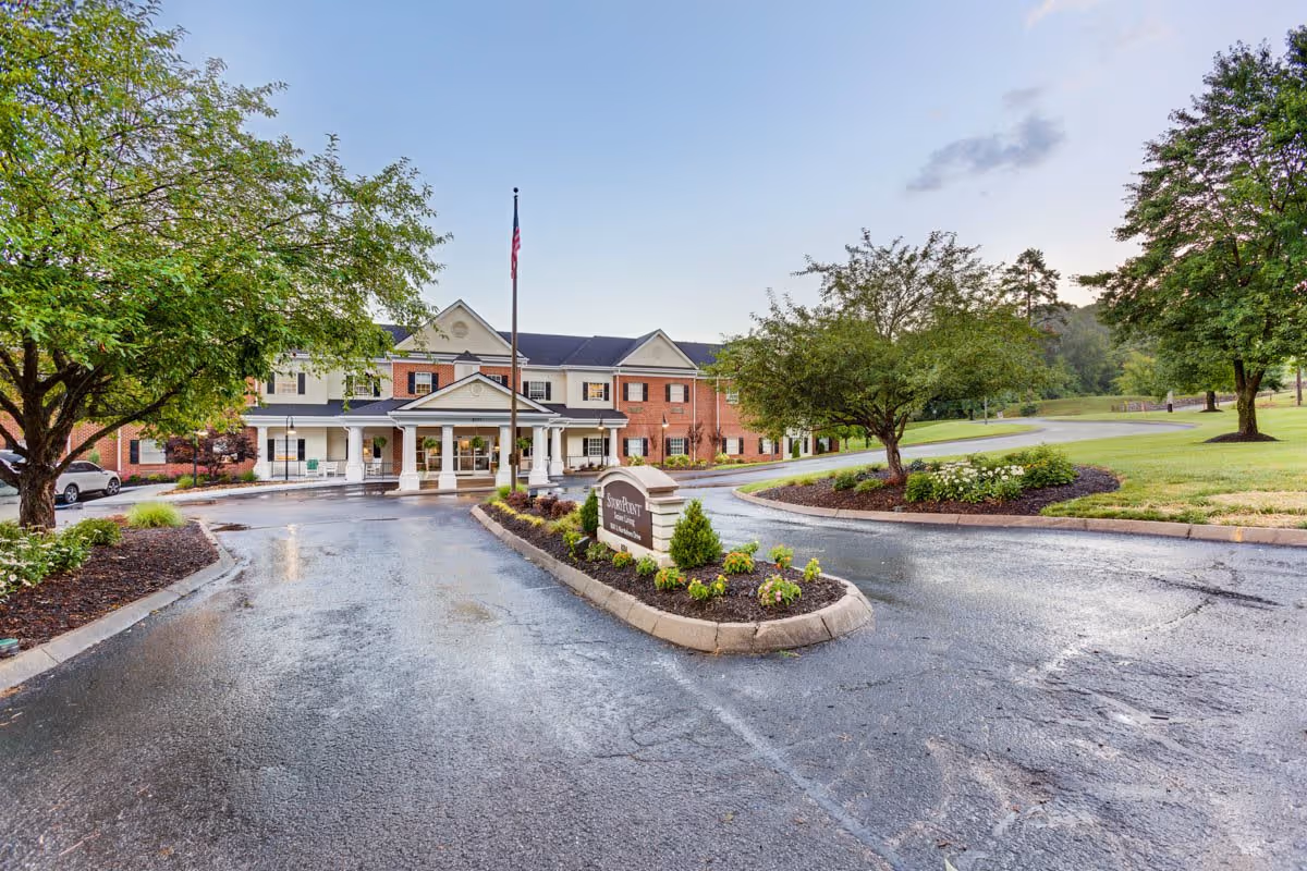 Exterior view of StoryPoint Knoxville West senior living facility with a driveway leading to the entrance, landscaped greenery, trees, and an American flag on a flagpole.