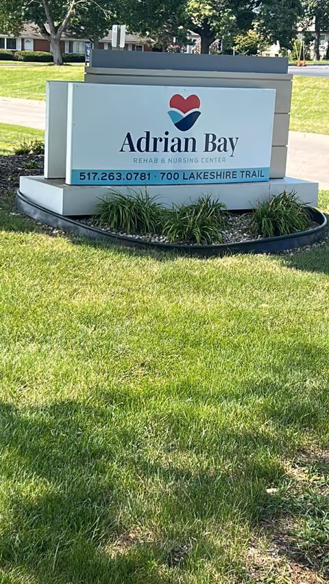 Outdoor view of a sign for Adrian Bay Rehab and Nursing Center, placed on a landscaped area with green grass and small plants. The sign includes a heart-shaped logo and contact information with a phone number and address.