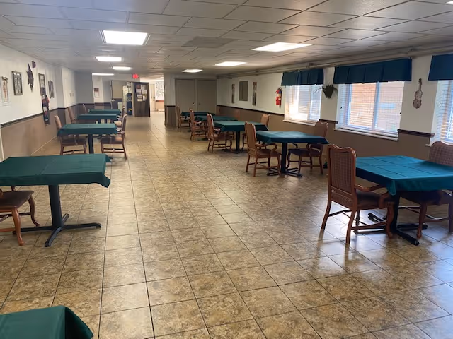 A long dining room with multiple tables covered in green tablecloths and wooden chairs arranged around them. The room has tiled flooring, white walls with brown wainscoting, and windows with green valances letting in natural light. The ceiling has recessed lighting and there are various decorations on the walls.