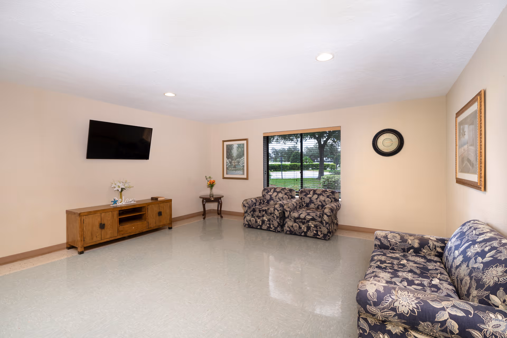 A simple living room with beige walls and a light-colored floor. The room contains a wooden TV stand with a flat-screen TV mounted above it, a small side table with a vase of flowers, two matching floral-patterned armchairs, and a floral-patterned sofa. There are framed pictures on the walls and a window showing greenery outside.