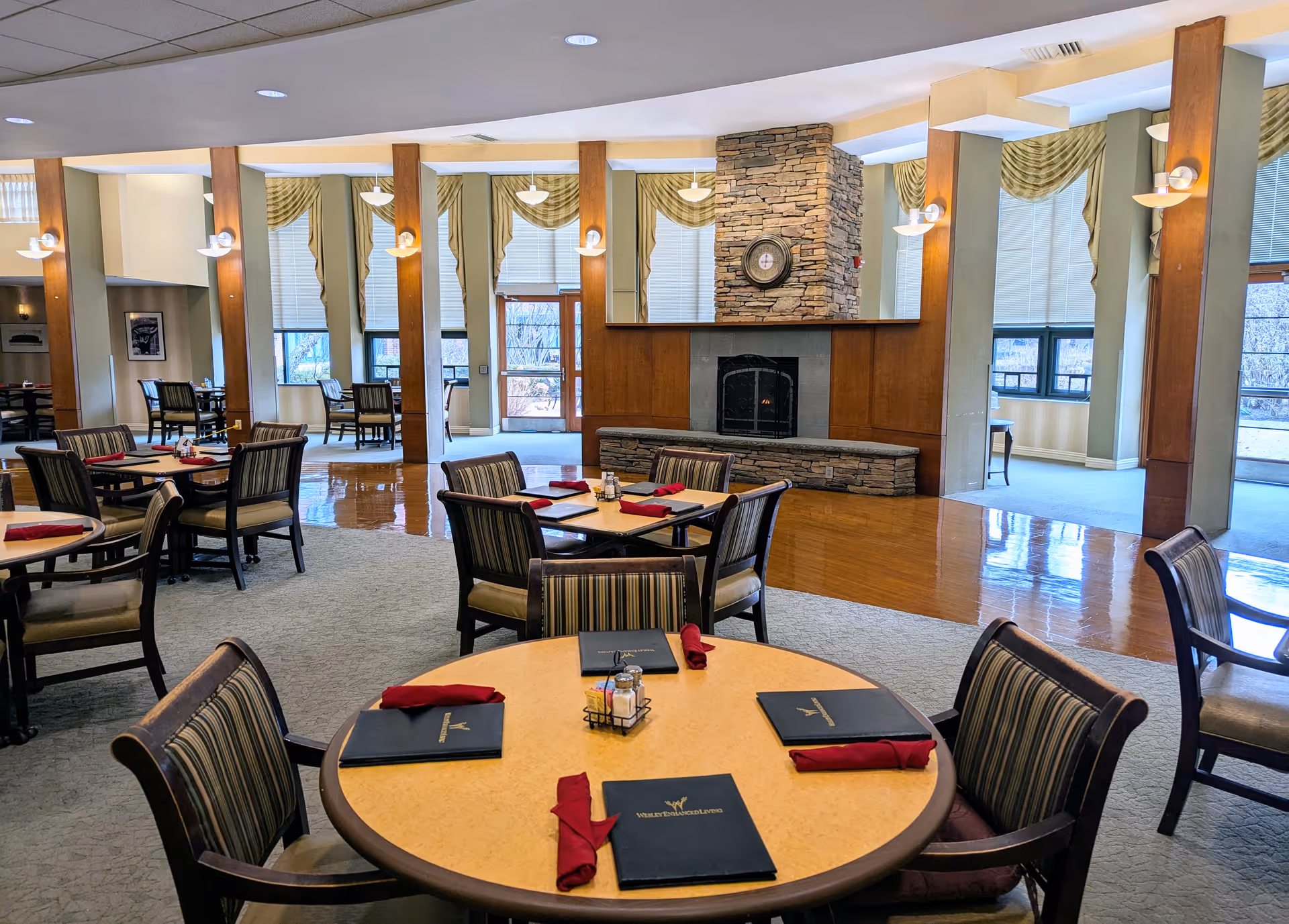Spacious senior dining room with round tables set with menus and napkins, chairs, and a central stone fireplace.