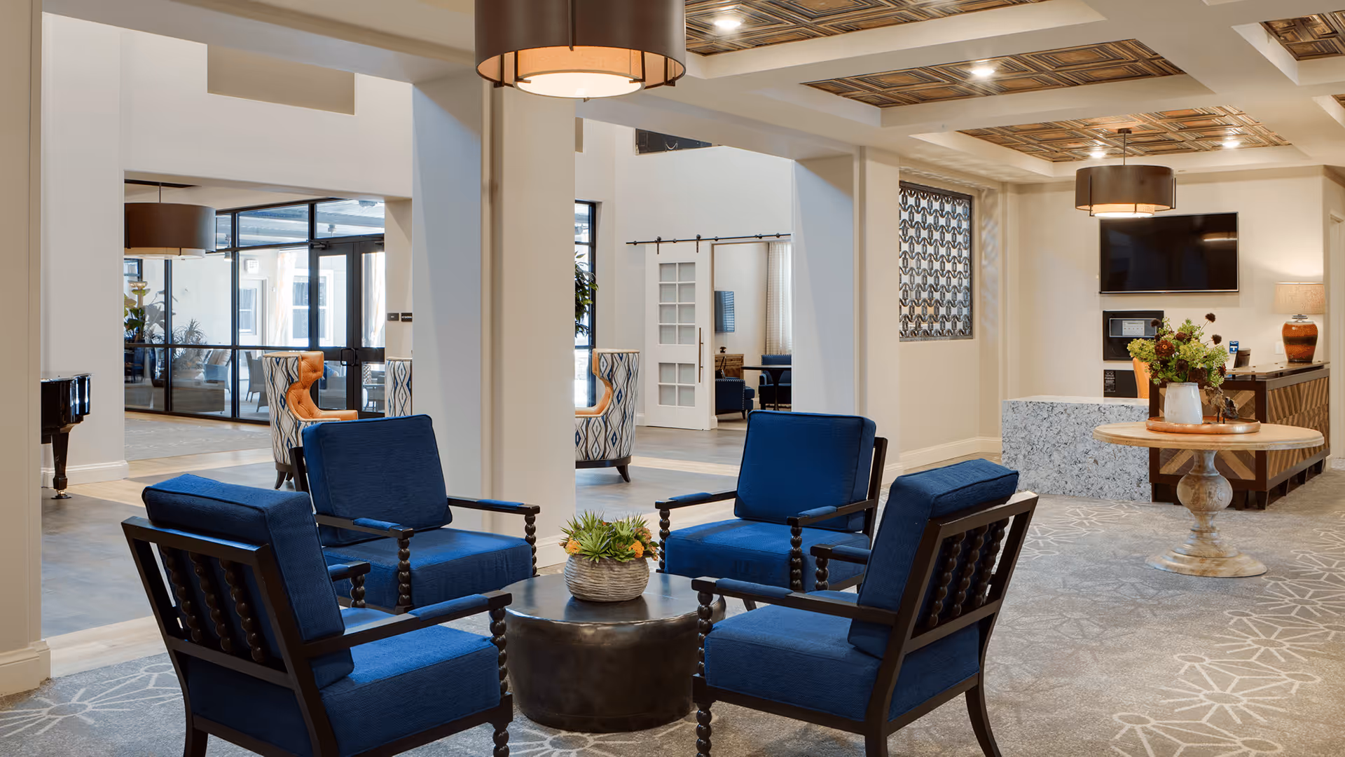 A spacious and well-lit common area with four blue cushioned armchairs arranged around a round black coffee table with a small plant centerpiece. The room features light-colored walls, a patterned carpet, and a decorative ceiling with recessed lighting. In the background, there is a round wooden table with a flower arrangement, a wall-mounted TV, and a reception desk area. Large windows and open doorways provide views into adjacent rooms with additional seating.