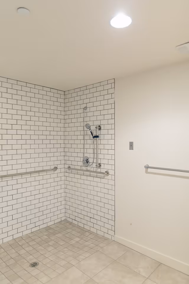 A spacious, accessible shower area with white subway tile walls and beige floor tiles. The shower features a handheld showerhead attached to a vertical grab bar, horizontal grab bars on two walls, and a ceiling light above.