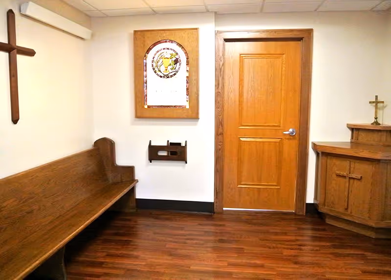 A small chapel or prayer room with wooden pew seating on the left, a wooden door in the center, and a wooden altar with a cross on the right. A wooden cross is mounted on the wall to the left, and a framed stained glass artwork is hung above a small wooden holder on the wall.