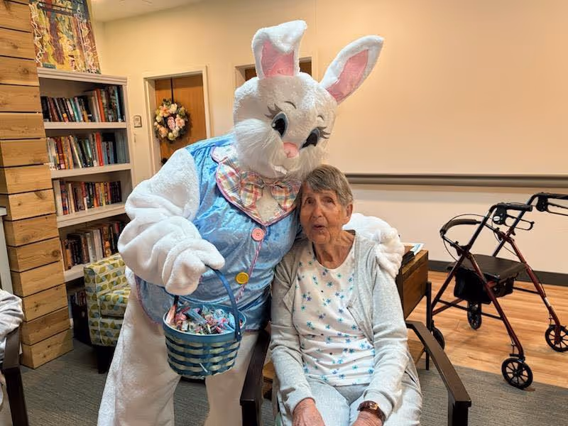 An elderly woman sitting in a chair inside a room with wooden flooring and a walker nearby. Next to her is a person dressed in a large white Easter Bunny costume wearing a blue vest and holding a basket of candy, with their arm around the woman. In the background, there is a bookshelf filled with books and a floral wreath hanging on a door.