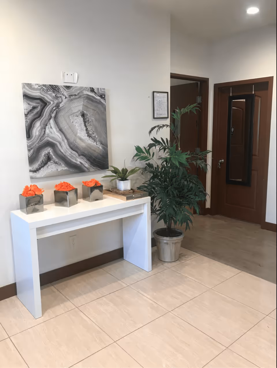 Interior corner of a facility with a white console table holding three small square vases with orange flowers and one small plant in a white pot. Above the table is a large abstract black and white wall art. To the right, there is a large potted green plant next to two brown doors, one with a full-length mirror attached. The floor is tiled and the walls are light-colored.