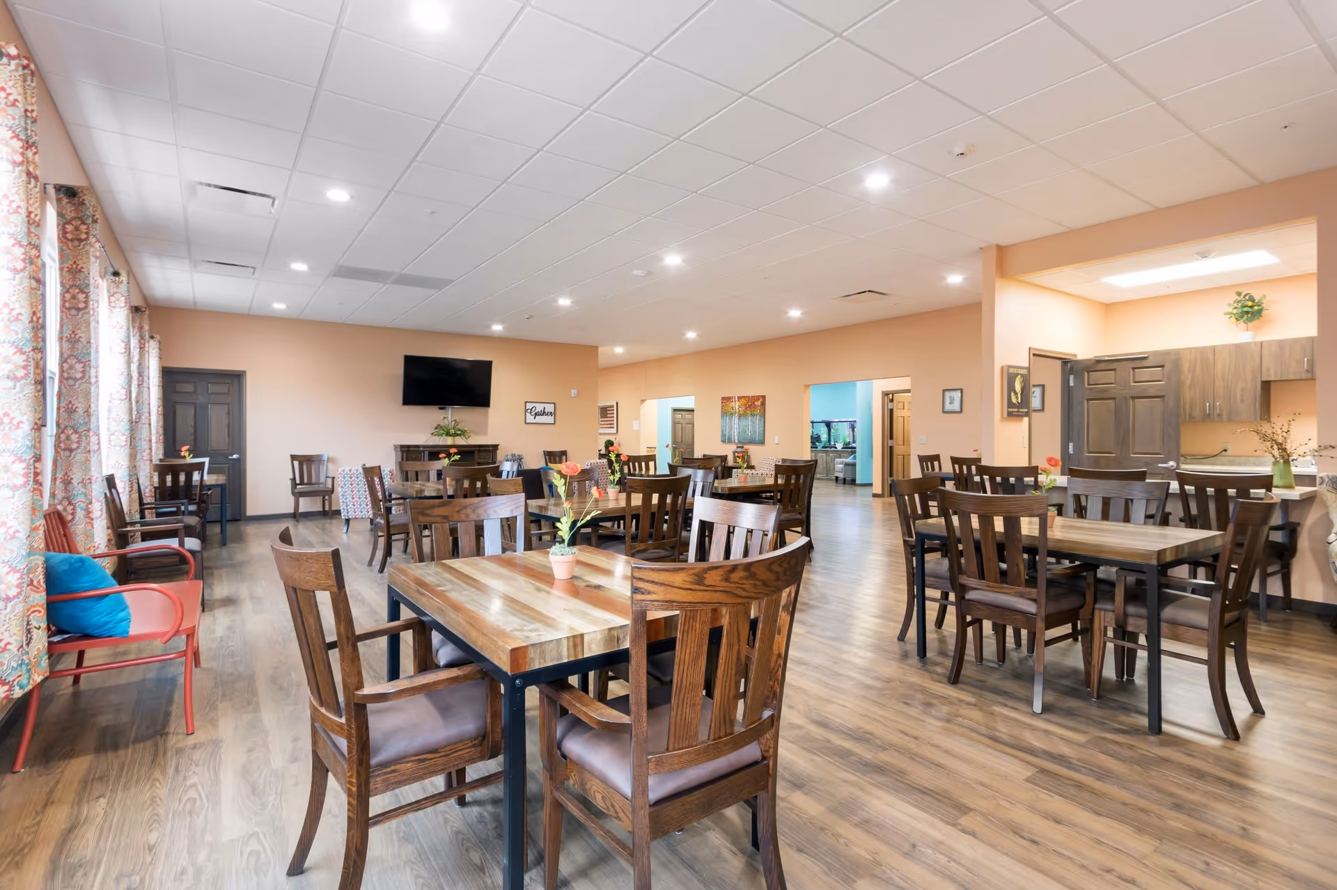 Bright communal dining and activity room with wooden tables and chairs, a TV on the far wall, and patterned curtains along the left windows.