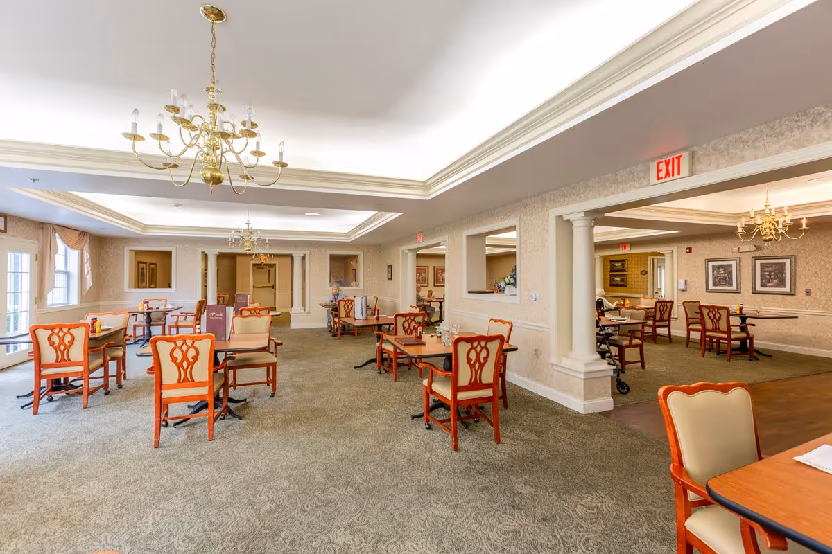 A spacious dining room in a senior living facility with multiple wooden tables and chairs arranged neatly. The room features elegant chandeliers, carpeted floors, large windows with drapes allowing natural light, and light-colored walls with decorative molding. There are exit signs visible above doorways and a few residents seated at tables in the background.