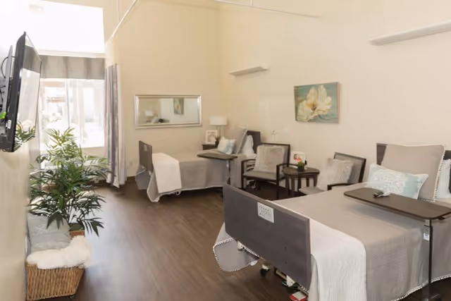 A bright and clean senior living room with two hospital-style beds, each with a beige bedspread and pillows. There are two chairs with a small table between them, a large wall mirror, a potted plant, and a wall-mounted TV. A window with curtains allows natural light to fill the room, and a floral painting hangs on the wall.