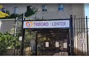 Entrance gate to Triboro Center with a sign displaying the facility's name above the gate. The gate is black metal with fencing on either side, and a building is visible in the background.