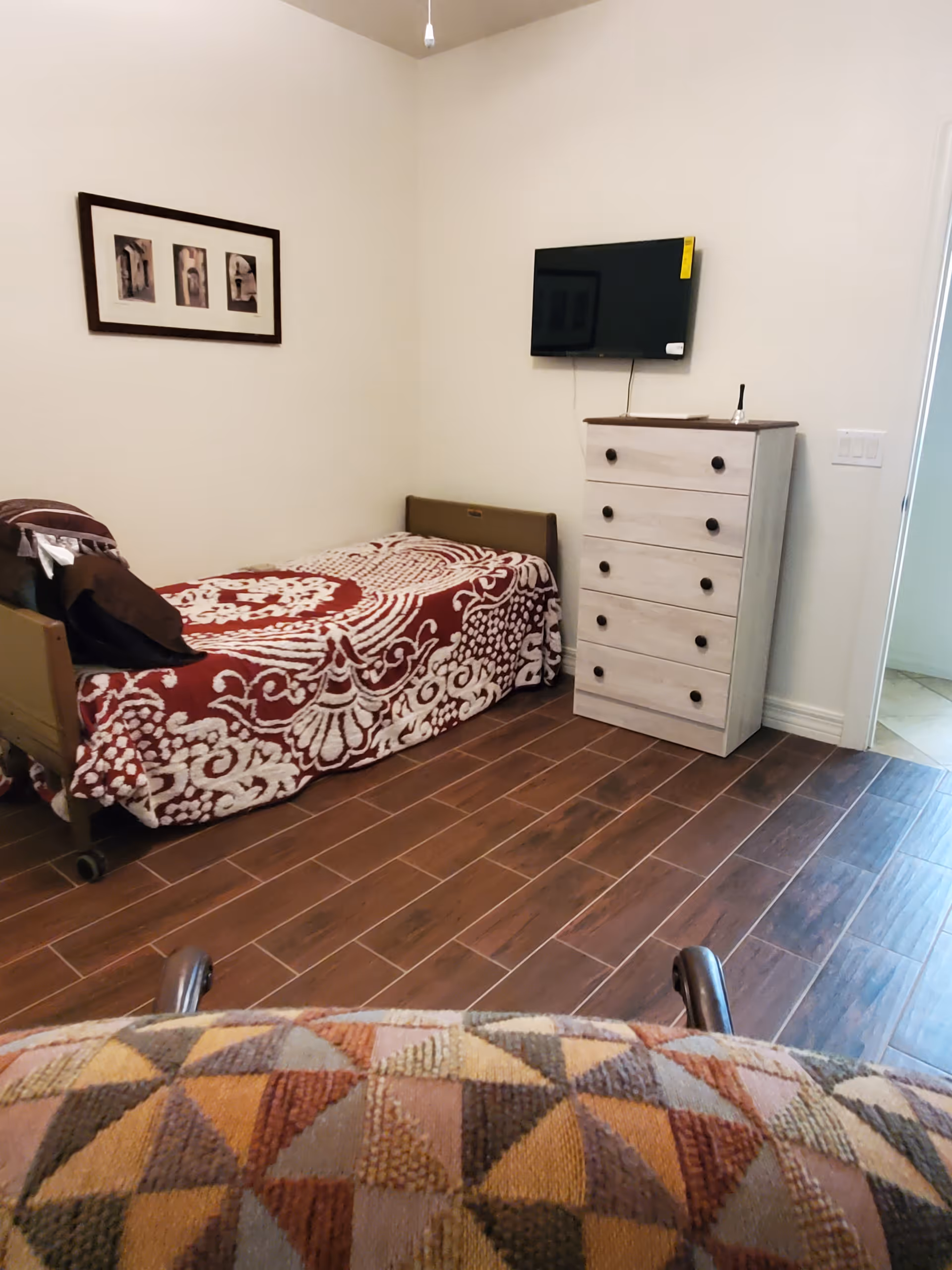 A small bedroom with a single bed covered in a red and white patterned blanket. There is a white chest of drawers with black knobs against the wall, and a flat-screen TV mounted above it. A framed picture with three images hangs on the wall above the bed. The floor is wooden, and part of a patterned chair is visible in the foreground.