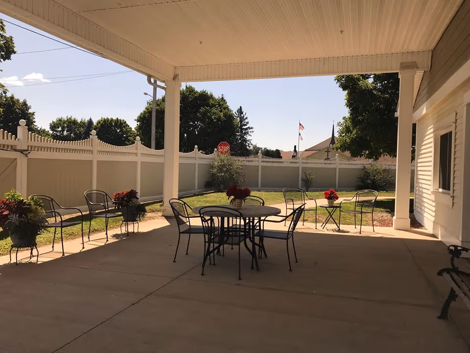 Covered outdoor patio area with metal tables and chairs, potted plants with red flowers on the tables, a white fence surrounding a grassy yard, trees in the background, and a stop sign visible beyond the fence.