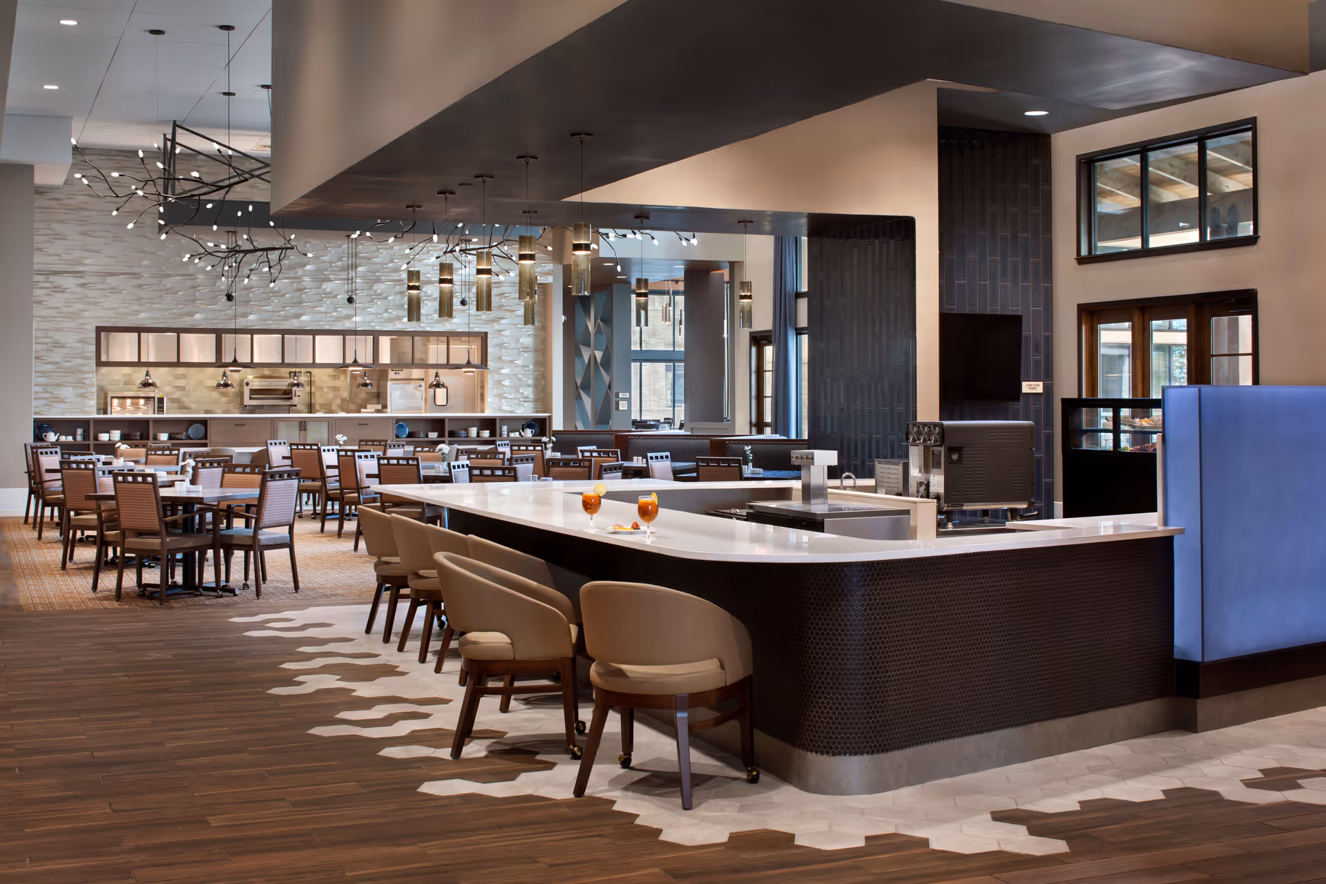 Modern dining area with multiple tables and chairs, a large curved bar with cushioned stools, pendant lighting, and large windows allowing natural light in.
