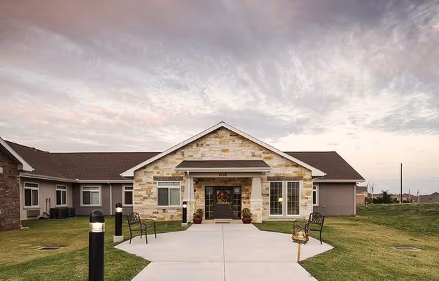 Front exterior view of a single-story building with a stone facade and a covered entrance. There are two benches on either side of the concrete walkway leading to the entrance, with grass lawns on both sides. The sky is partly cloudy during sunset or sunrise.