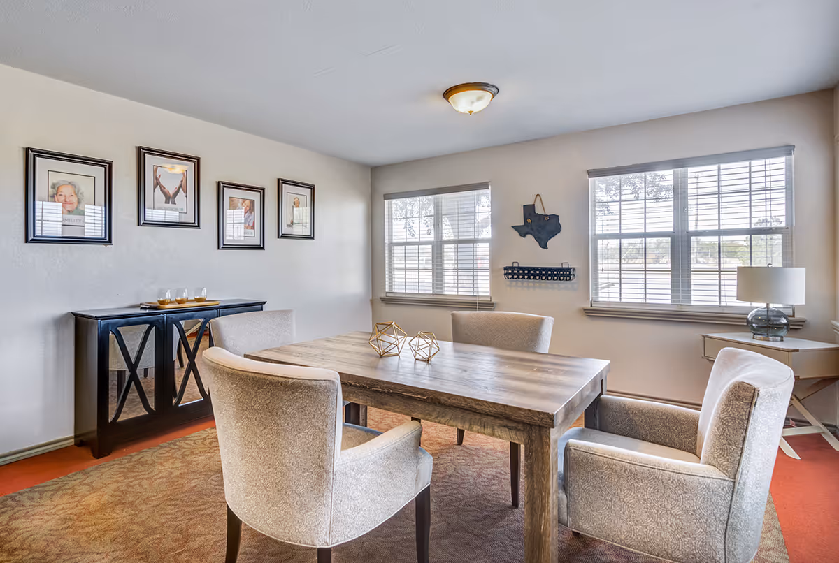A bright room with a wooden table surrounded by four upholstered chairs. The room has two large windows with white blinds, a small side table with a lamp, and a black cabinet against the wall with framed pictures above it. The walls are light-colored and the floor is carpeted.