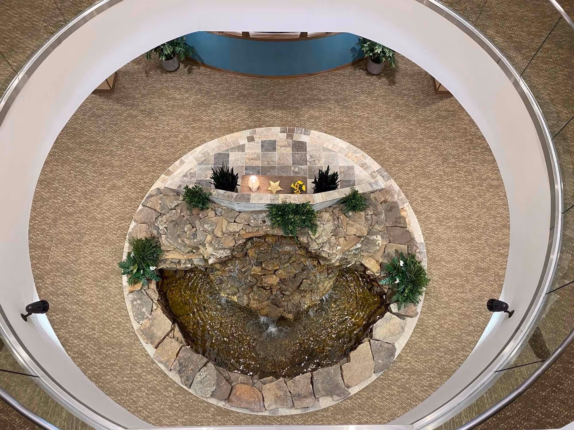 Top-down view of a circular indoor water feature with rocks, plants, and a small waterfall, surrounded by a carpeted floor and a curved white railing.