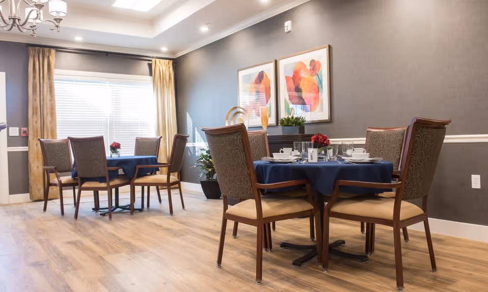 A dining area with two round tables covered with dark blue tablecloths, each surrounded by four wooden chairs with cushioned seats and backs. The tables are set with white dishes, cups, and silverware, and decorated with small flower arrangements. The room has light wood flooring, gray walls with white trim, two colorful abstract paintings, large windows with beige curtains, and a chandelier hanging from the ceiling.