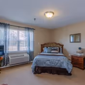 Well-lit bedroom with a double bed, nightstand, window with curtains, and a wall air-conditioning unit.
