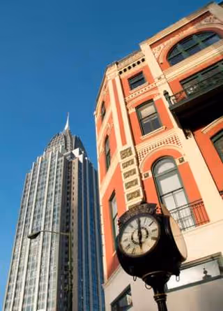 A red brick building with arched windows towers beside a modern glass skyscraper under blue sky