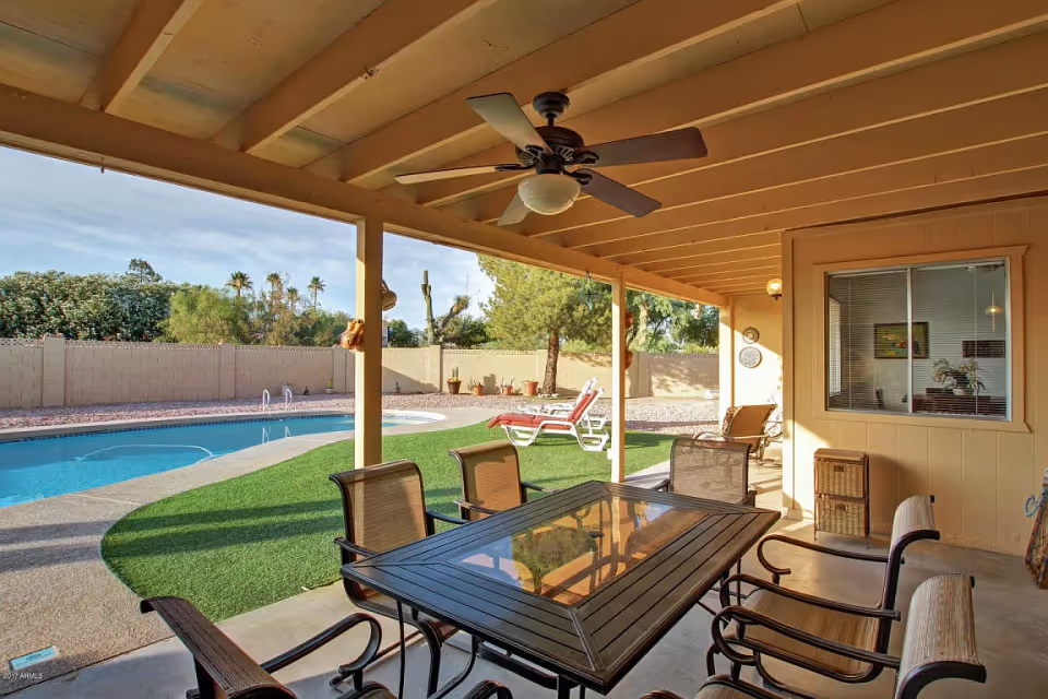 Covered outdoor patio area with a ceiling fan, a glass-top table, and six chairs. Beyond the patio is a swimming pool surrounded by a concrete deck and artificial grass. Two lounge chairs with red cushions are positioned near the pool. The area is enclosed by a beige brick wall with trees and shrubs visible outside.