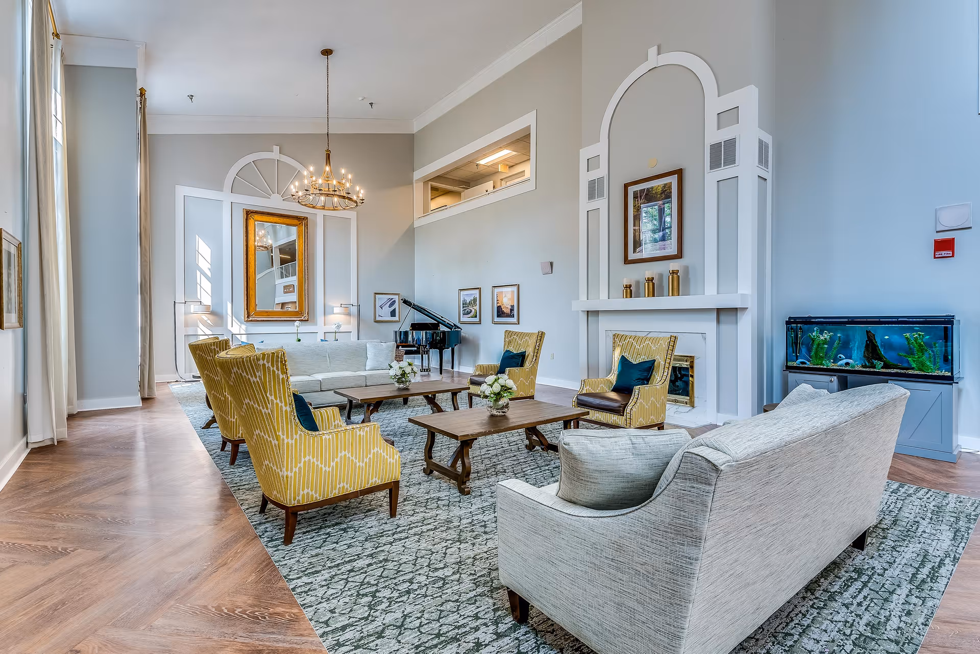 Bright communal living room with sofas, yellow armchairs, a chandelier, fireplace, grand piano, and an aquarium.