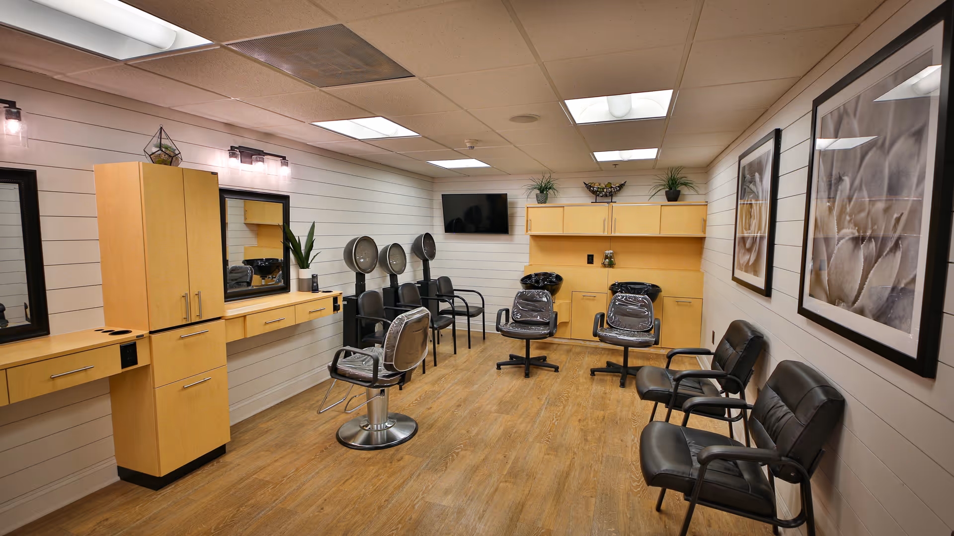 Interior of a salon room with wooden flooring and light-colored walls. The room features salon chairs, hair drying stations, mirrors, cabinets, and framed floral artwork on the walls. There are also some potted plants and a wall-mounted TV.