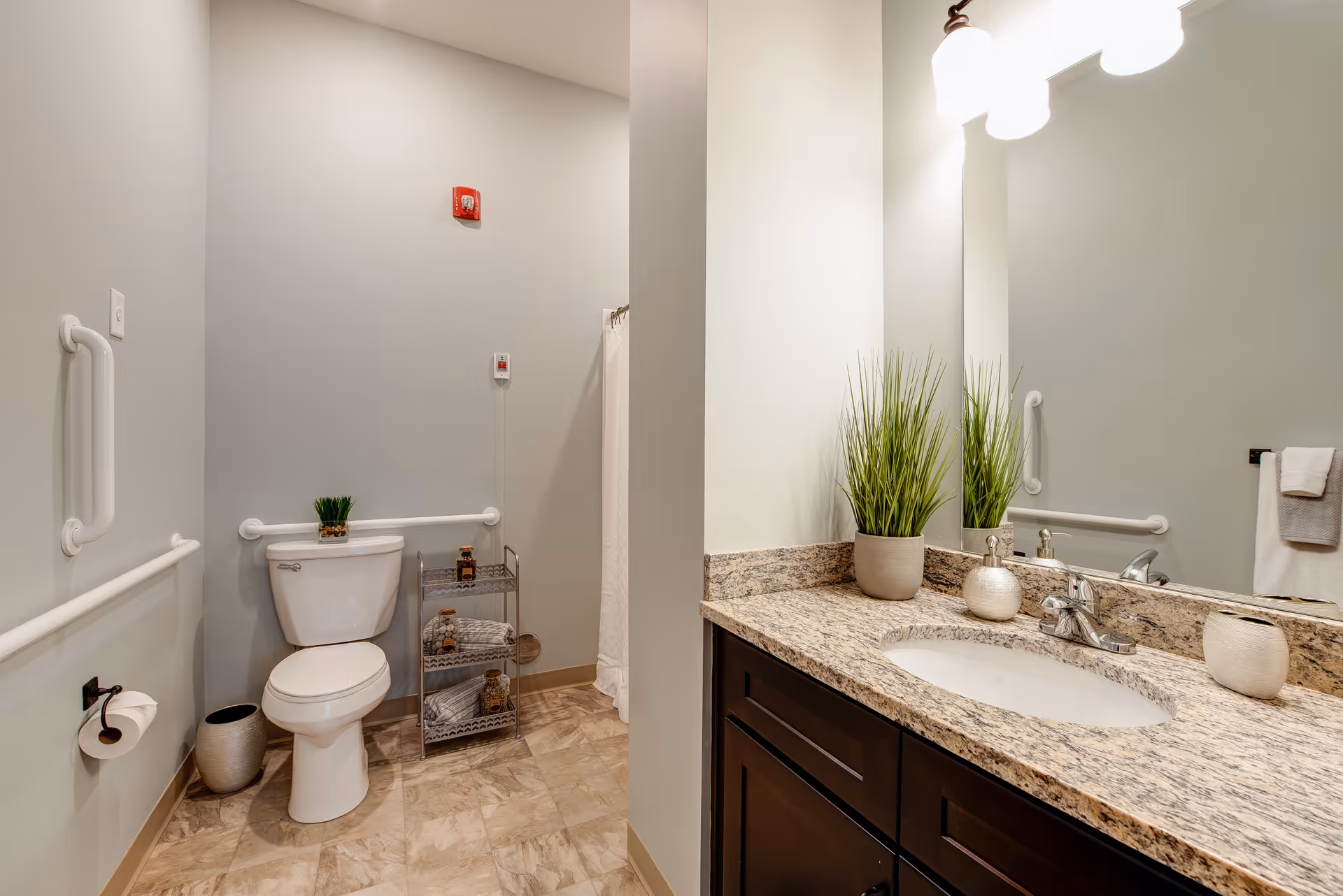 A clean and modern bathroom with a toilet, grab bars on the walls, a small metal shelf holding towels and decorative items, and a granite countertop with a sink, faucet, and potted plant. A large mirror and wall-mounted lights are above the sink.