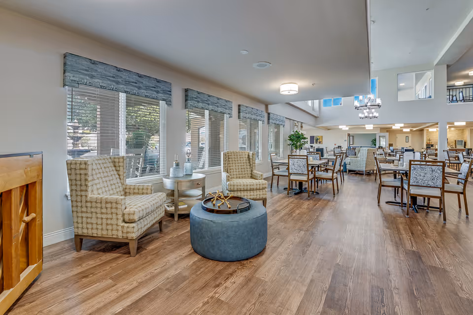 Spacious senior living common dining area with tables, chairs, lounge seating, and large windows.